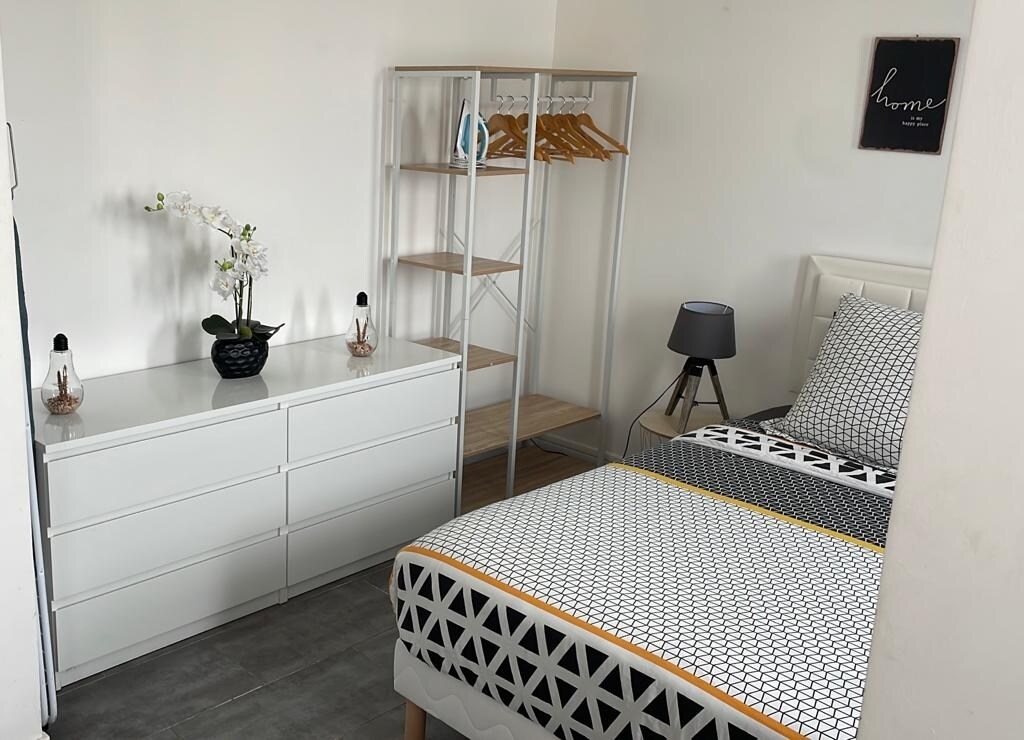 A neat sleeping area features a spacious bed with patterned bedding. A white dresser and a modern clothing rack provide storage. A potted orchid and table lamp add decorative touches, while a framed sign reading 'home' is displayed on the wall.