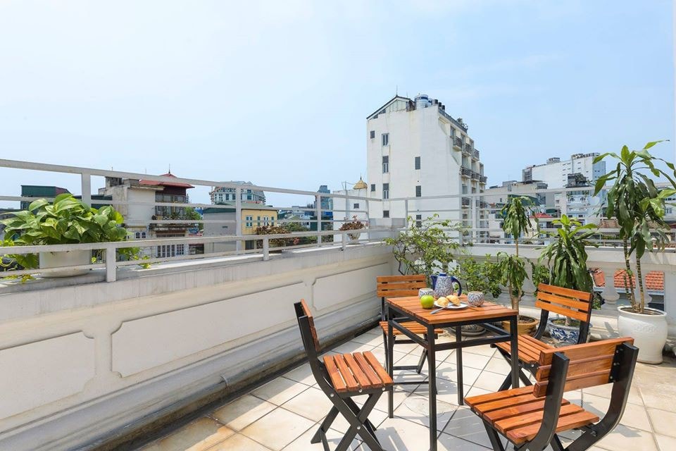 The spacious balcony features a wooden table surrounded by four chairs, ideal for outdoor dining or relaxation. Potted plants add greenery to the area, which offers views of the surrounding buildings and cityscape under a clear sky.