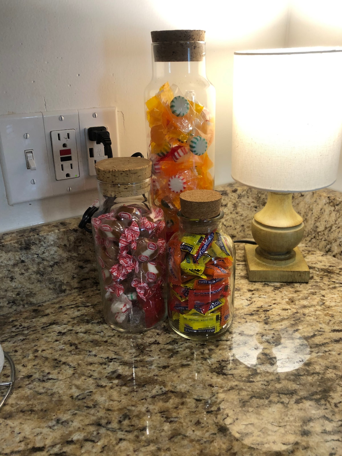 Three glass jars filled with assorted candies are displayed on a granite countertop. The jars feature cork lids, with one jar containing wrapped pink candies, another filled with colorful hard candies, and the third showcasing various pieces of taffy. A lamp is positioned nearby.