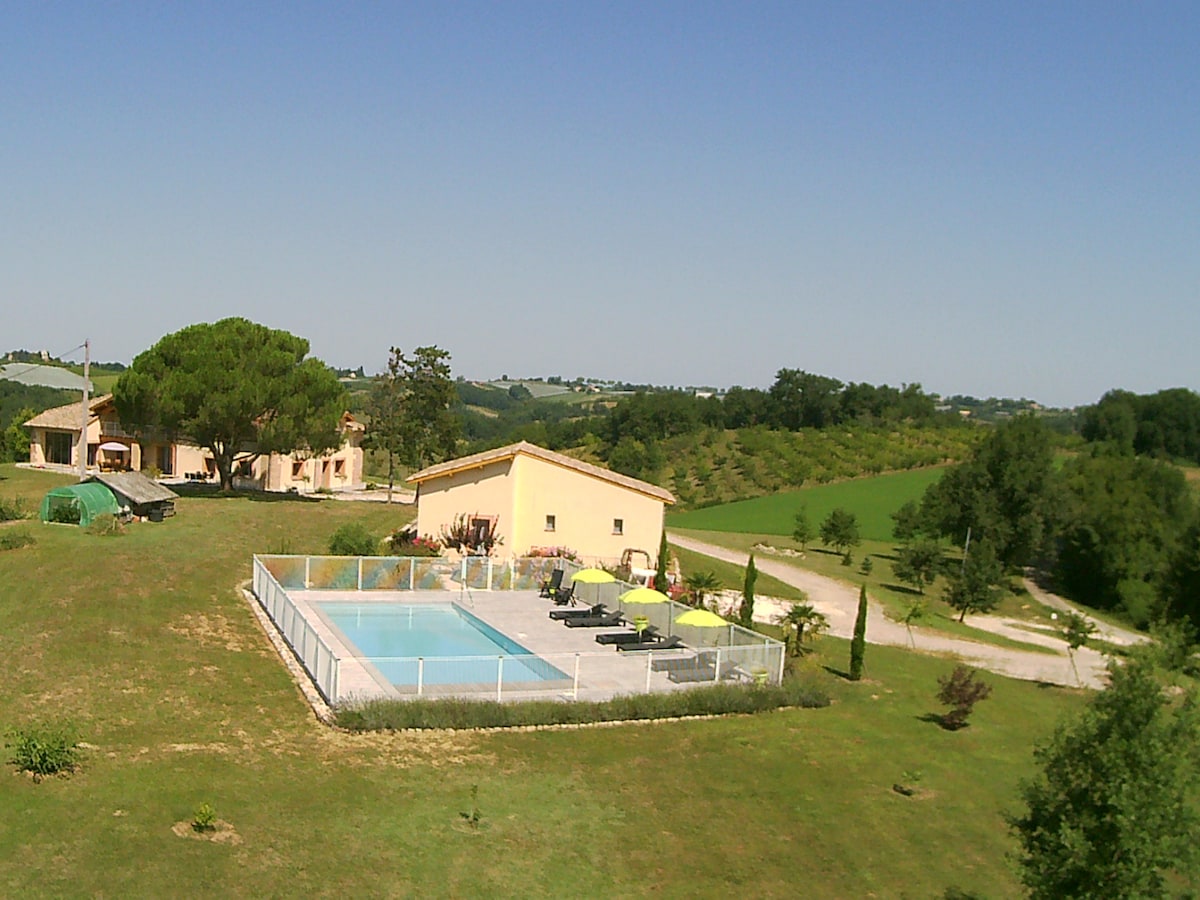 The image showcases a serene landscape with a fenced swimming pool in the foreground. The pool is surrounded by lounge chairs and a patio area. In the background, the rustic architecture of the house is visible, nestled among lush greenery and rolling hills under a clear blue sky.