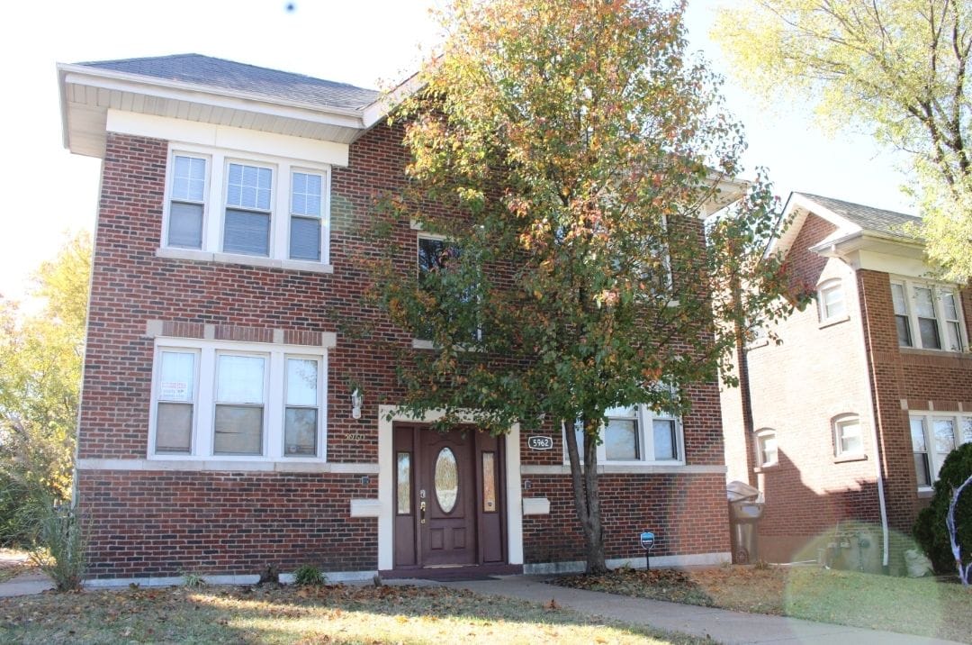 A three-story brick building features multiple windows and a prominent entrance with double doors. The surrounding area showcases well-maintained landscaping, including a mature tree with autumn foliage. Sunlight enhances the building's warm brick tones, creating a welcoming exterior.