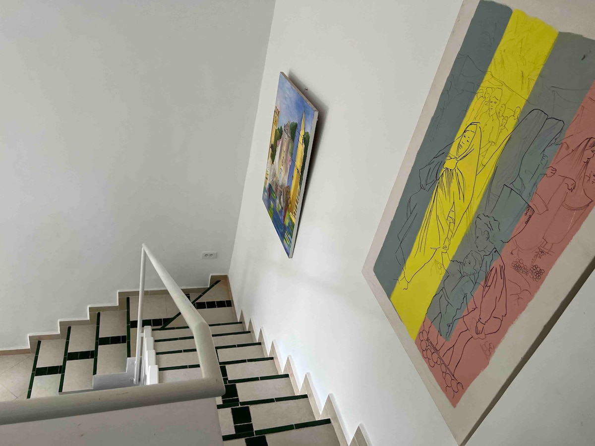 A modern staircase is featured, with vibrant artwork displayed on the white walls. The floor is laid with black and white tiles, leading downward. A handrail provides support along the stairs, creating a functional and aesthetically pleasing transition between levels.