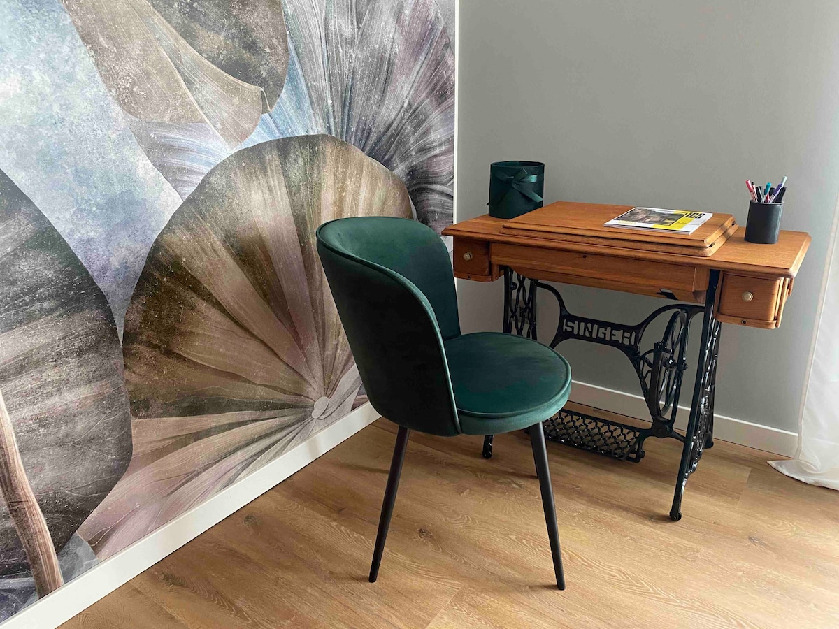 A workspace is presented with a wooden desk featuring an industrial-style base and a green chair. A wall mural of overlapping leaves adds a natural aesthetic, while a small speaker and colorful stationary are also visible on the desk.