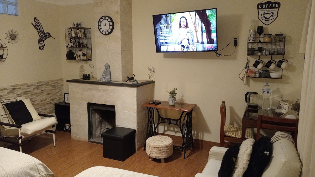 A cozy living area is highlighted by a functional fireplace and a mounted television, which is positioned on a light-colored wall. Decorative elements include wall art and a clock. Equipped furniture includes a small dining table, chairs, and a comfortable seating arrangement.