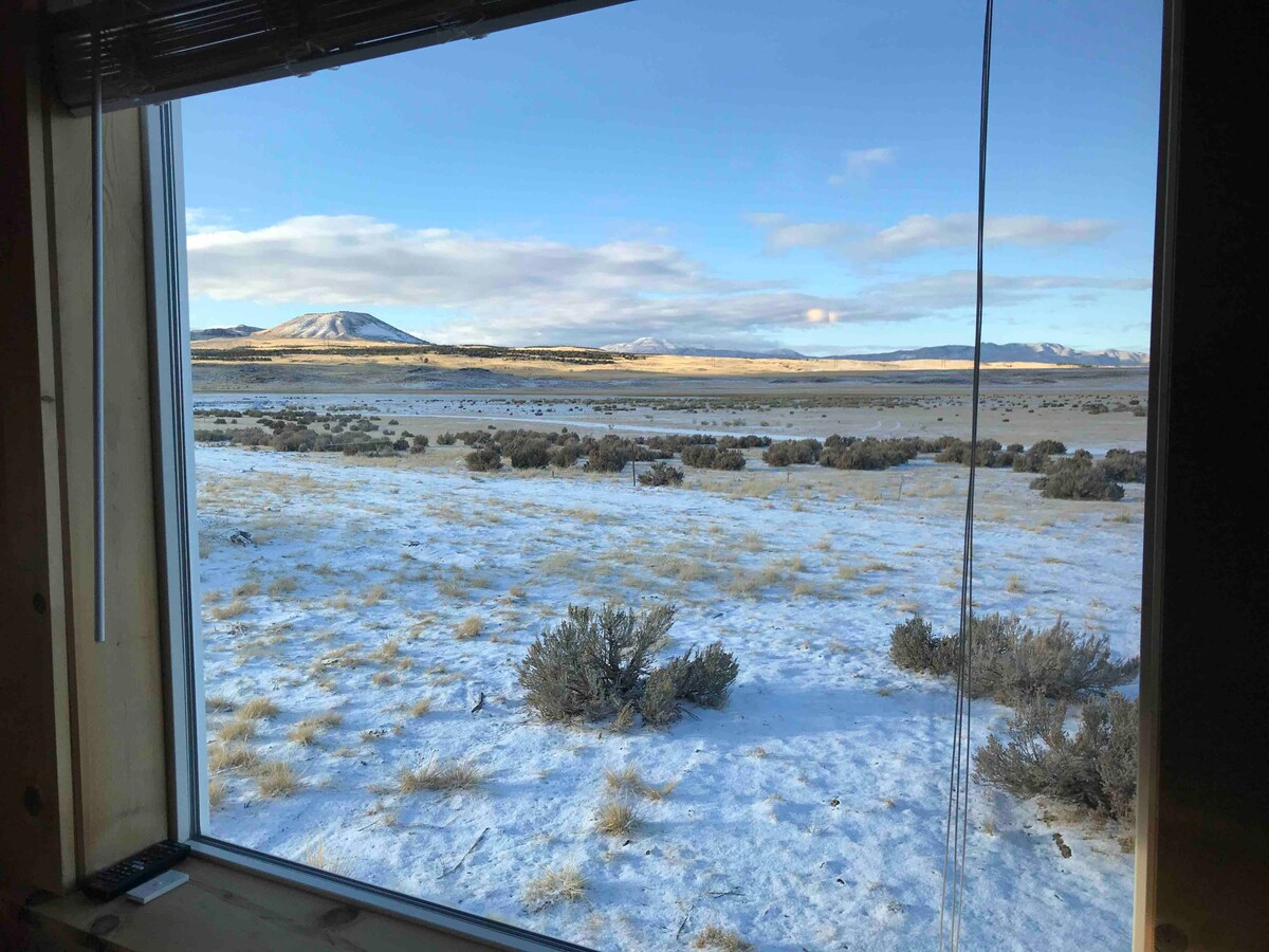 A wide window frame displays a serene winter landscape, featuring rolling hills dusted with snow and sparse vegetation. The light blue sky is visible, contrasting with the sunlit terrain. Mountains are seen distantly against a calm horizon, enhancing the tranquil atmosphere.