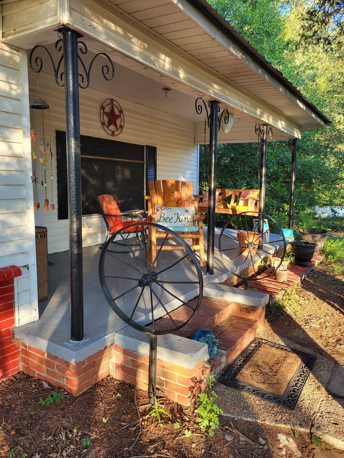 A welcoming front porch features two wooden rocking chairs, enhanced by decorative wagon wheels. A rustic star emblem is visible on the wall. The entryway is framed by lush greenery, creating a tranquil outdoor atmosphere.