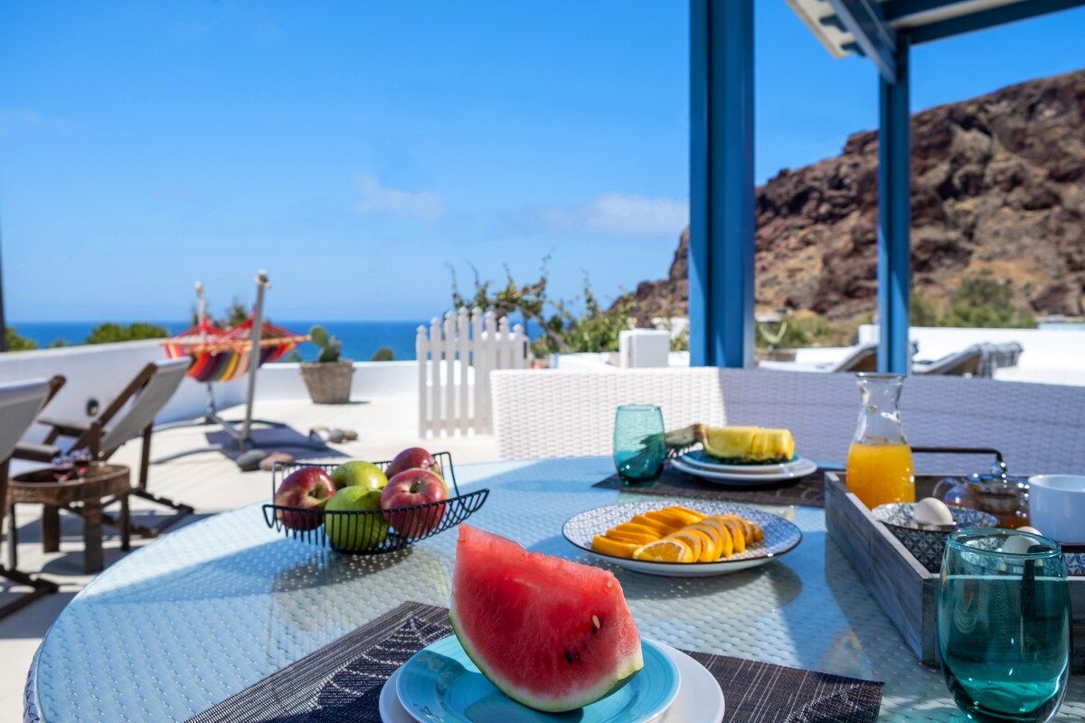 An outdoor dining area is set for breakfast, featuring a glass table adorned with plates of fresh fruit, including watermelon, apples, and oranges. A jug of orange juice and glasses are ready for service, with a scenic view of the Aegean Sea visible in the background.