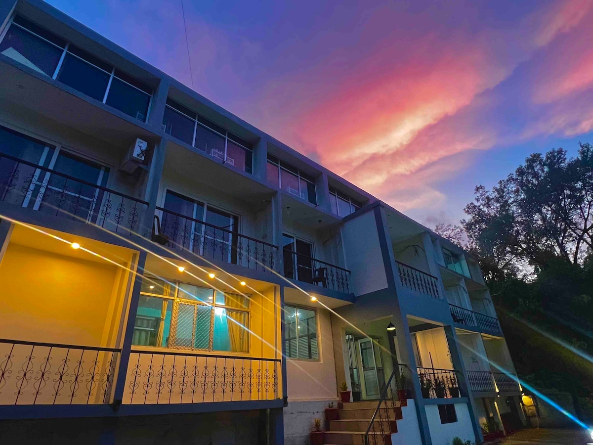 A modern building features multiple balconies with black railings. Large windows reflect the colorful sunset, creating a serene atmosphere. Soft lighting highlights the entrance, while lush trees frame the property, emphasizing the connection to nature.