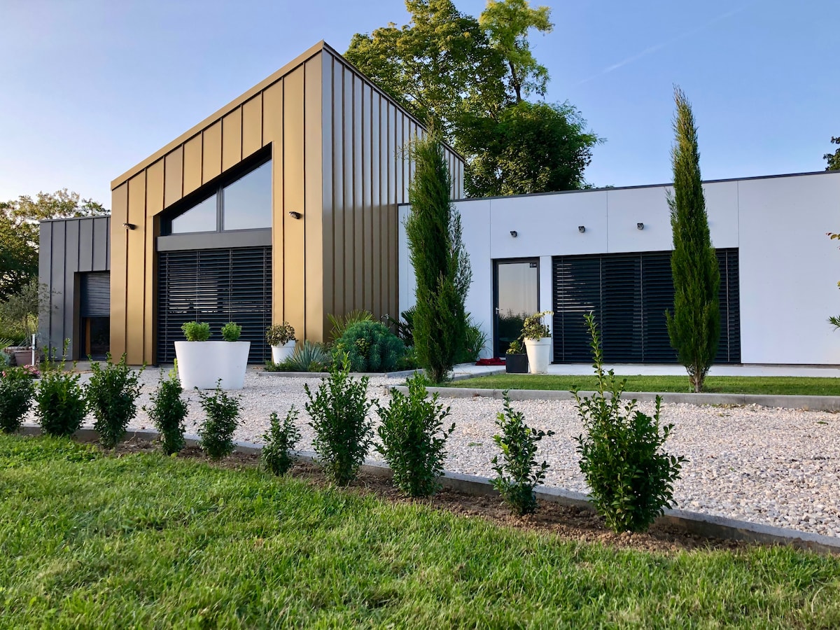 A modern architectural home is presented, featuring a striking gold and white exterior. Tall, narrow windows are arranged symmetrically, and landscaped greenery borders the gravel pathway. Neatly trimmed shrubs and planters emphasize the clean lines of the design, creating a visually appealing entrance.