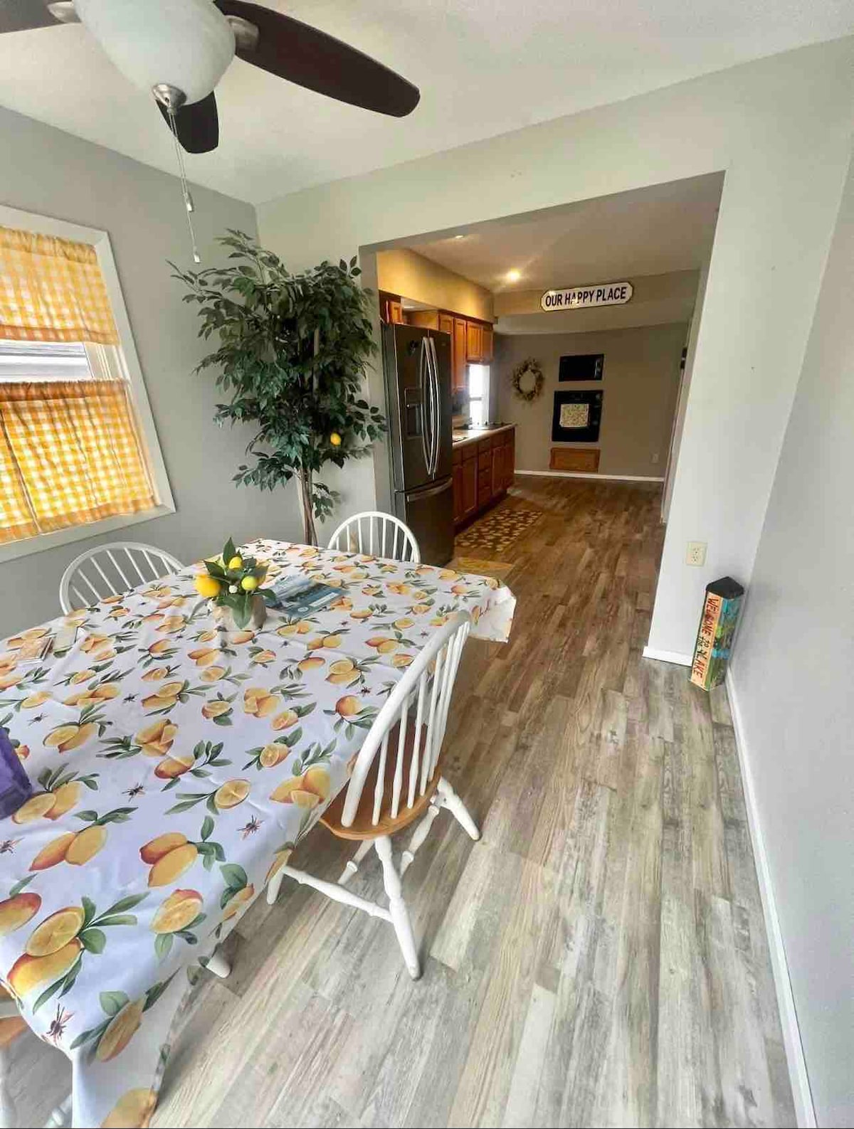 A dining area is shown, featuring a table covered with a tablecloth adorned with a floral pattern. White chairs surround the table, and a potted plant is positioned in the corner. The kitchen area is visible in the background, showcasing cabinets and appliances.