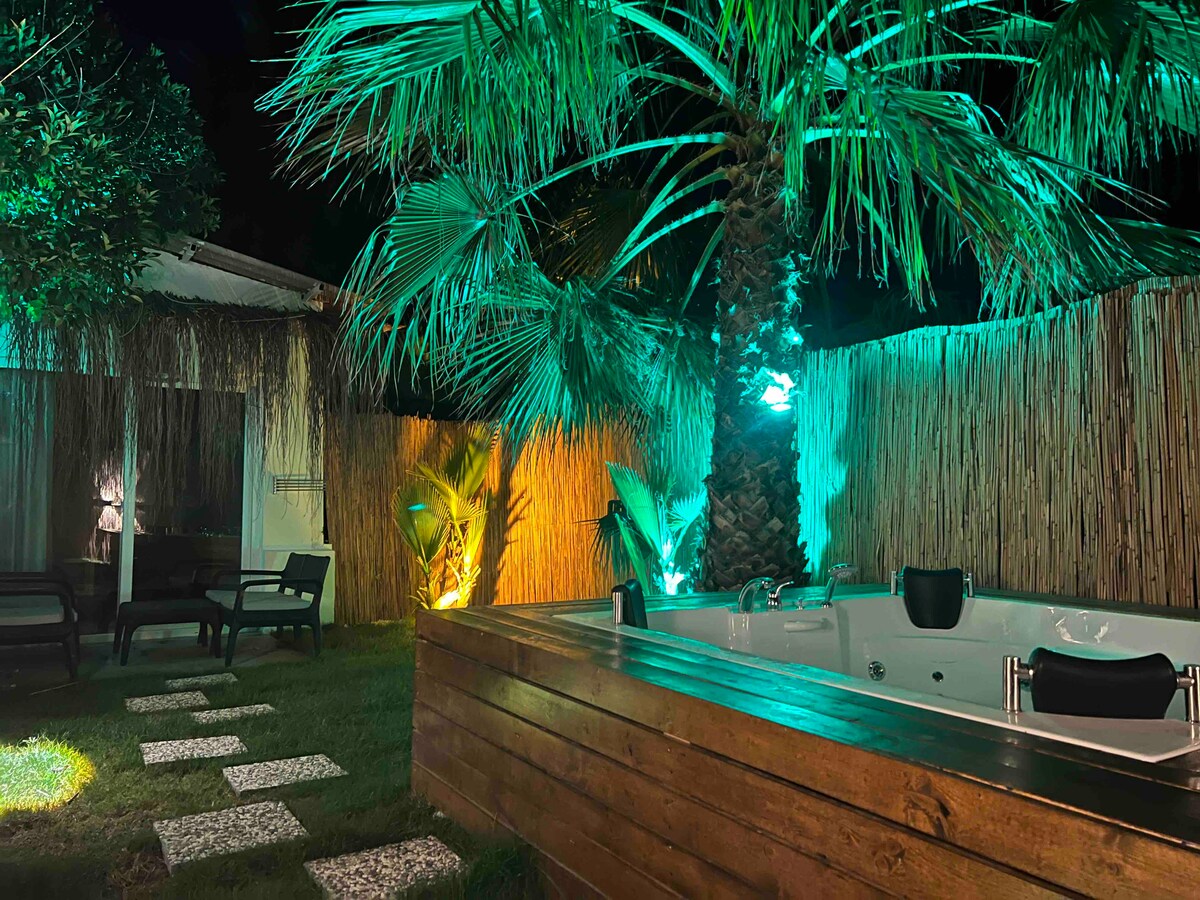 A private garden area features a spacious hot tub surrounded by lush greenery and palm trees. Soft lighting accentuates a tranquil atmosphere, while neatly arranged stepping stones lead through the well-kept lawn. Comfortable seating is provided nearby, enhancing the relaxation space.