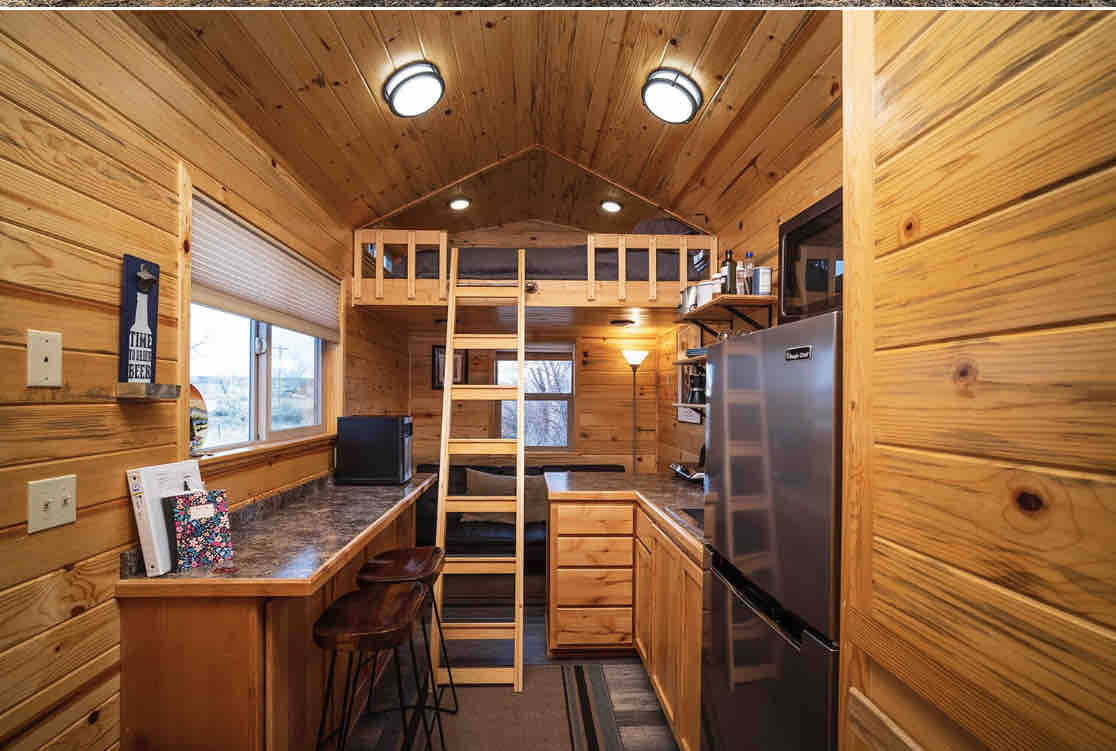 The interior showcases warm wood paneling with a loft area accessible by a sturdy ladder. The kitchen features stainless steel appliances, a bar counter with two stools, and a view through a window. A computer workstation is positioned beside the seating area.