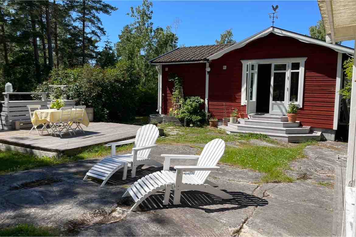 A red guest house is nestled among greenery, featuring light-colored steps leading to a welcoming entrance. Two white lounge chairs are positioned on a stone patio, overlooking a wooden deck with a dining table and chairs set for outdoor meals.