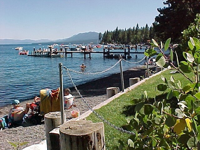 A scenic view of a pebbly beach along a lake is featured, with boats gently bobbing on the water. A wooden pier extends into the lake, while several individuals enjoy the sun. Lush green trees frame the area, enhancing the peaceful setting.