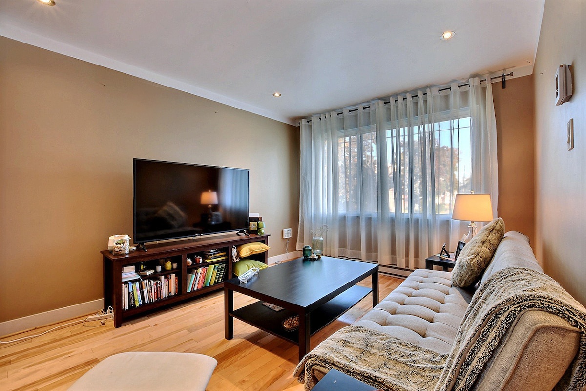 A cozy living room features a light-finished wooden floor and a large window draped with sheer curtains, allowing natural light to fill the space. A flat-screen television is mounted on the wall, and a low coffee table sits in front of a comfortable sofa adorned with decorative cushions.