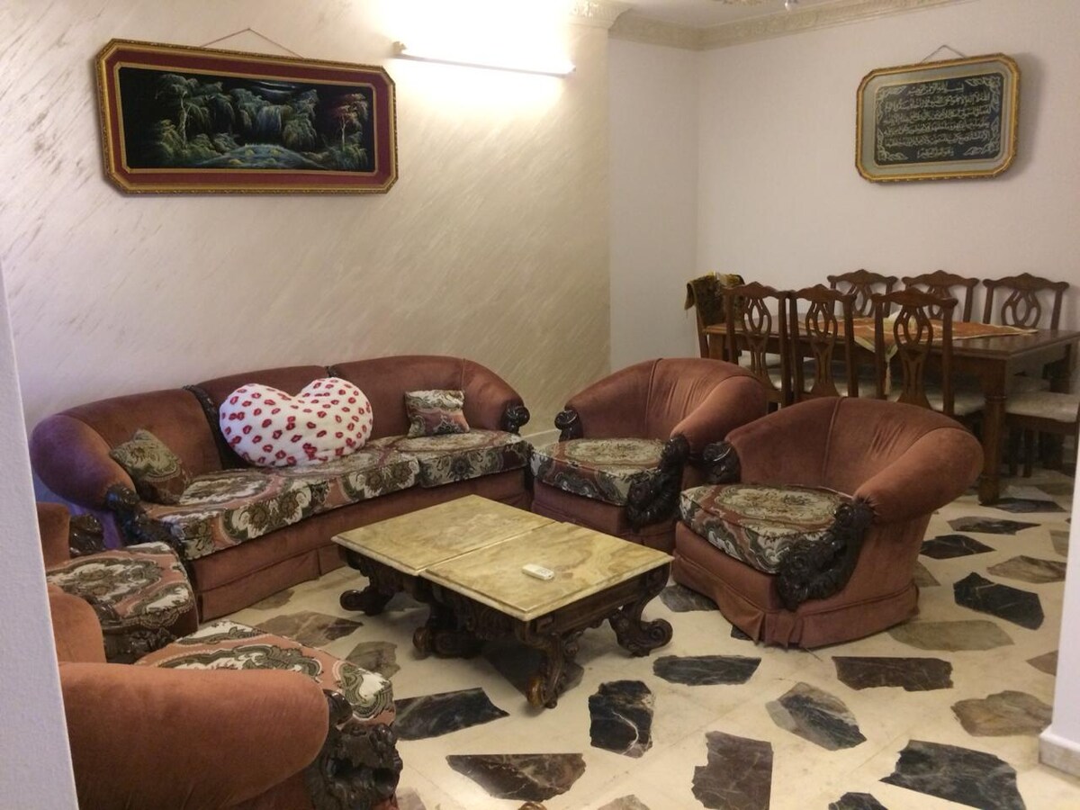 A lounge area features a heart-shaped pillow on a richly upholstered sofa, surrounded by matching armchairs. A stone coffee table sits at the center, while a wooden dining table and chairs are visible in the background, creating a cozy gathering space.