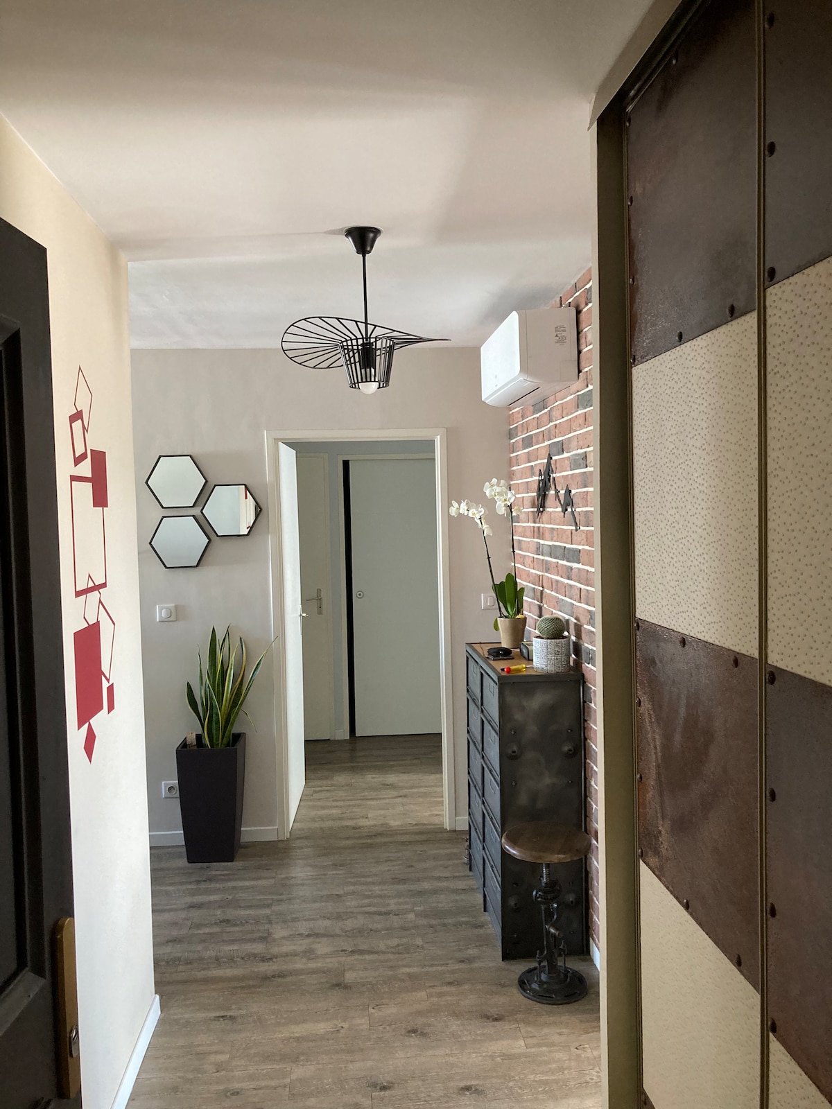 A hallway is presented with a blend of modern and industrial design elements. Geometric mirrors adorn the walls, alongside a decorative light fixture. A potted plant adds a touch of greenery, while a dark dresser and a stool create functional accents in the space.