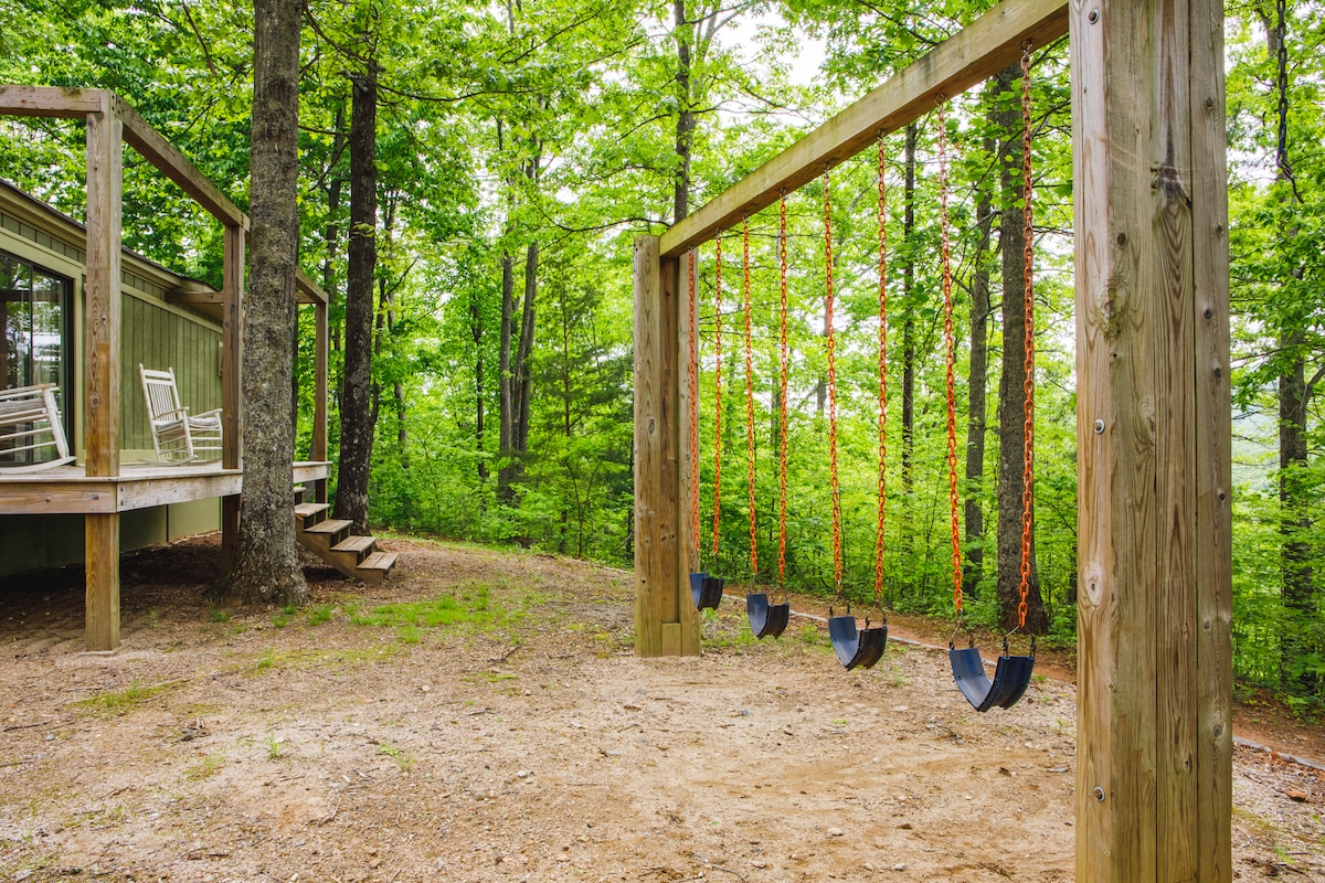 A wooded outdoor area features a set of swings hanging from sturdy wooden frames. Nearby, a deck is accessible via steps, providing a seating area with rocking chairs surrounded by lush greenery.