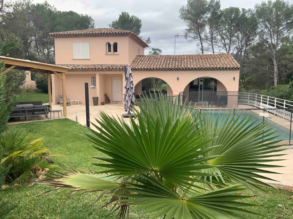 A villa is surrounded by lush greenery, featuring a spacious terrace with a pool and outdoor seating area. The building showcases a classic Mediterranean style with arched openings and a tiled roof. A palm plant adds a vibrant touch in the foreground.
