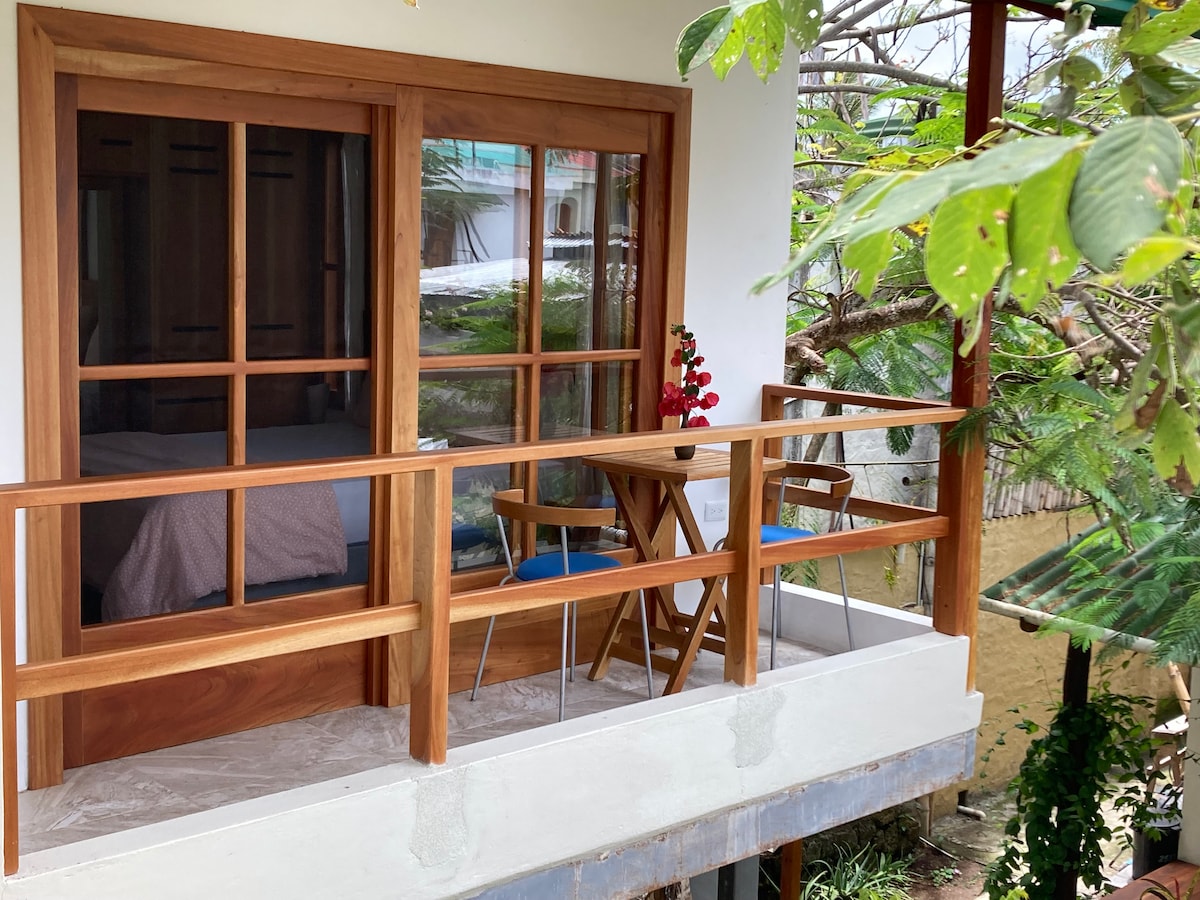 A private balcony features wooden railings and outdoor seating with two chairs and a small table. Lush greenery surrounds the area, providing a serene view. Large glass doors invite light into the room, softly framing the sleeping area visible through the window.