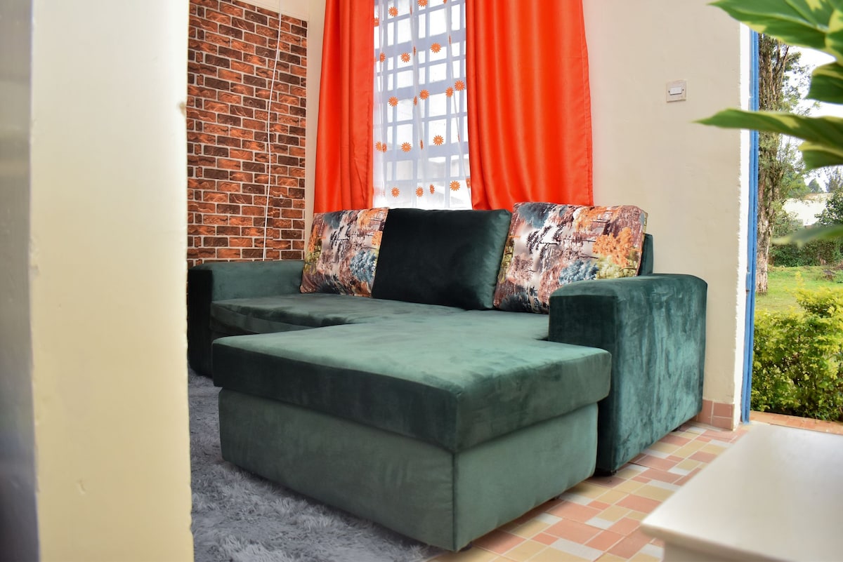 A comfortable green sectional sofa is positioned against a brick-patterned wall. Bright orange curtains frame a window, allowing natural light to illuminate the space. Soft gray carpeting is present on the floor, enhancing the cozy appearance of the seating area.