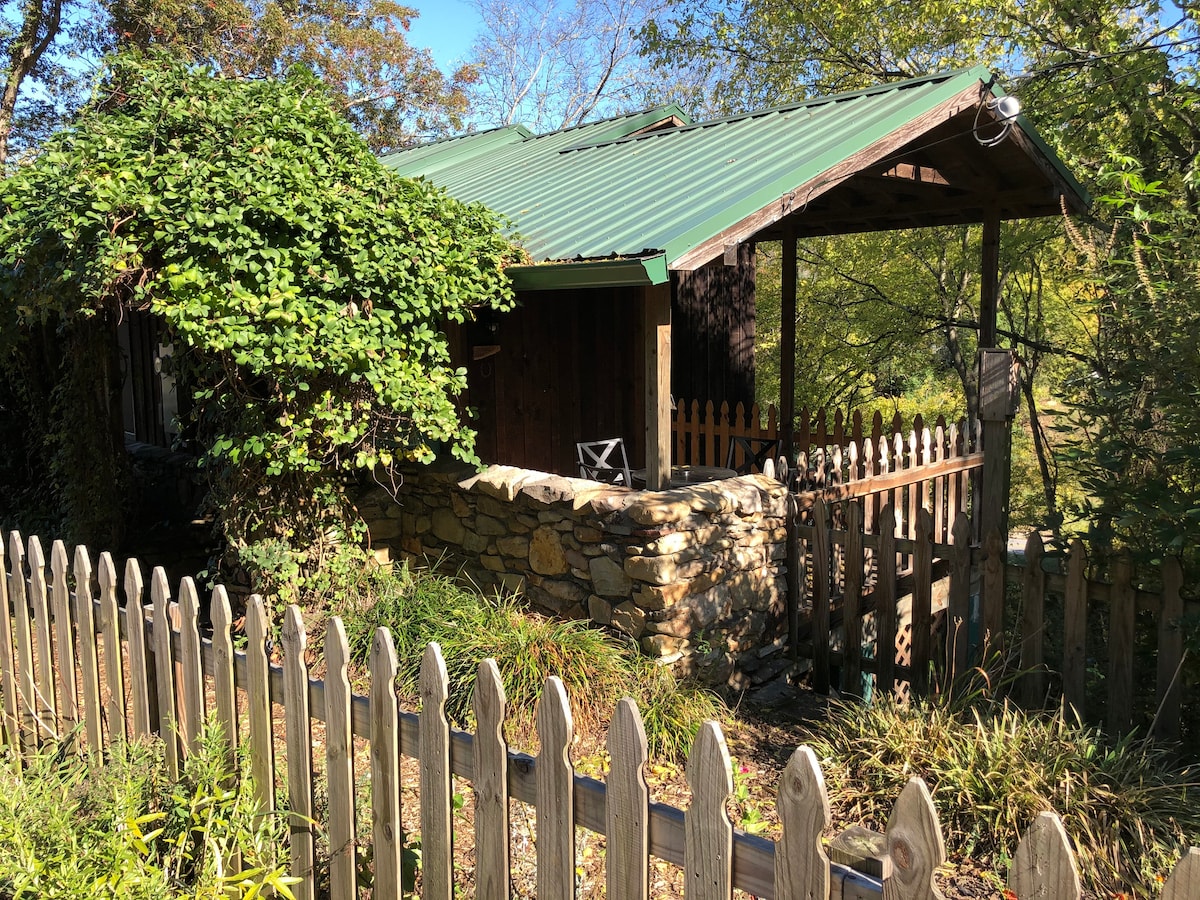 The cabin's exterior showcases a rustic stone wall and a green metal roof, surrounded by lush greenery. A small wooden chair sits on a stone porch, with a picket fence enclosing the space. Natural light filters through the trees, enhancing the inviting entrance.