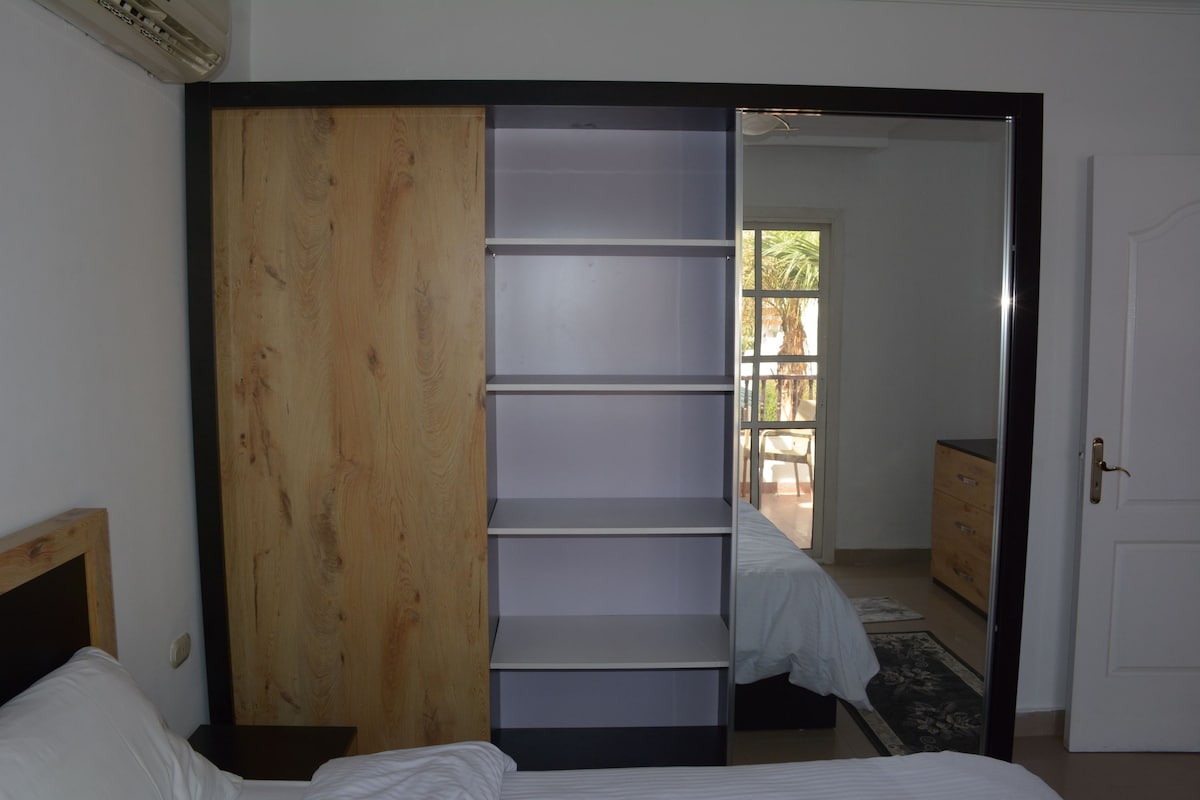 A sliding mirrored wardrobe is seen, with wooden and light-colored shelves. The reflection reveals a glimpse of the terrace and palm trees outside, enhancing the sense of space. Warm tones of the wooden furniture are visible, contributing to a calming environment.