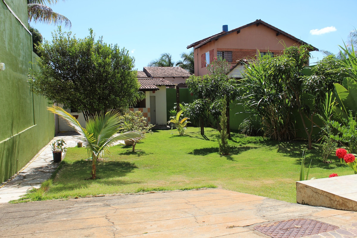 A green outdoor space features a well-maintained lawn, complemented by various shrubs and trees. The sun casts a bright light over the area, enhancing the inviting atmosphere. A pathway is visible leading towards the house, with a glimpse of adjacent buildings in the background.