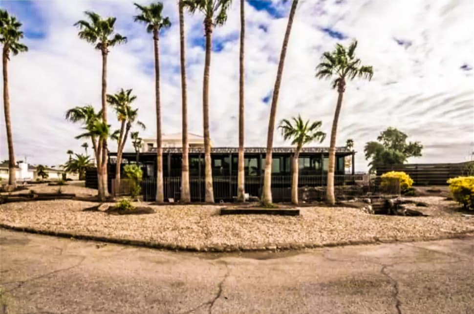 A modern home is framed by a row of tall palm trees, creating a tropical ambiance. The property features landscaped gravel areas with a variety of shrubs and plants. The sky above is partly cloudy, casting a soft light over the exterior.