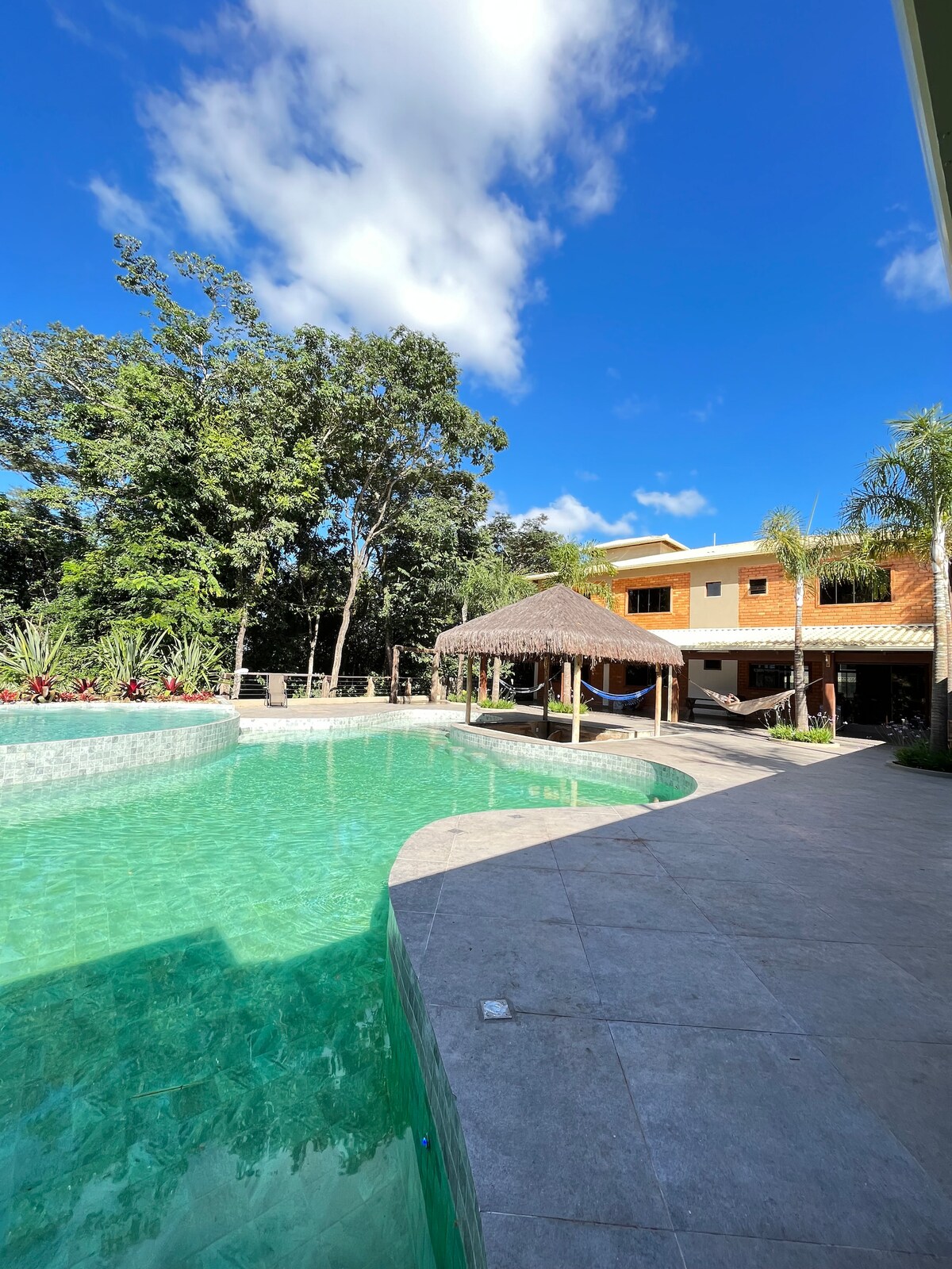 A luxurious pool area is surrounded by lush greenery, featuring a spacious pool with clear blue water. A thatched-roof gazebo provides shaded seating, while the exterior of the house displays a harmonious blend of warm colors against the vivid sky.