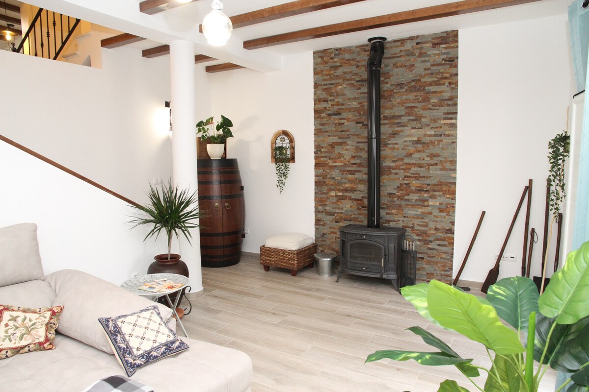 A cozy living area features a light beige sofa and a decorative pillow. A stone accent wall provides a rustic touch, complemented by a wood-burning stove. A plant adds a touch of greenery, while a barrel and woven storage basket enhance the space's charm.
