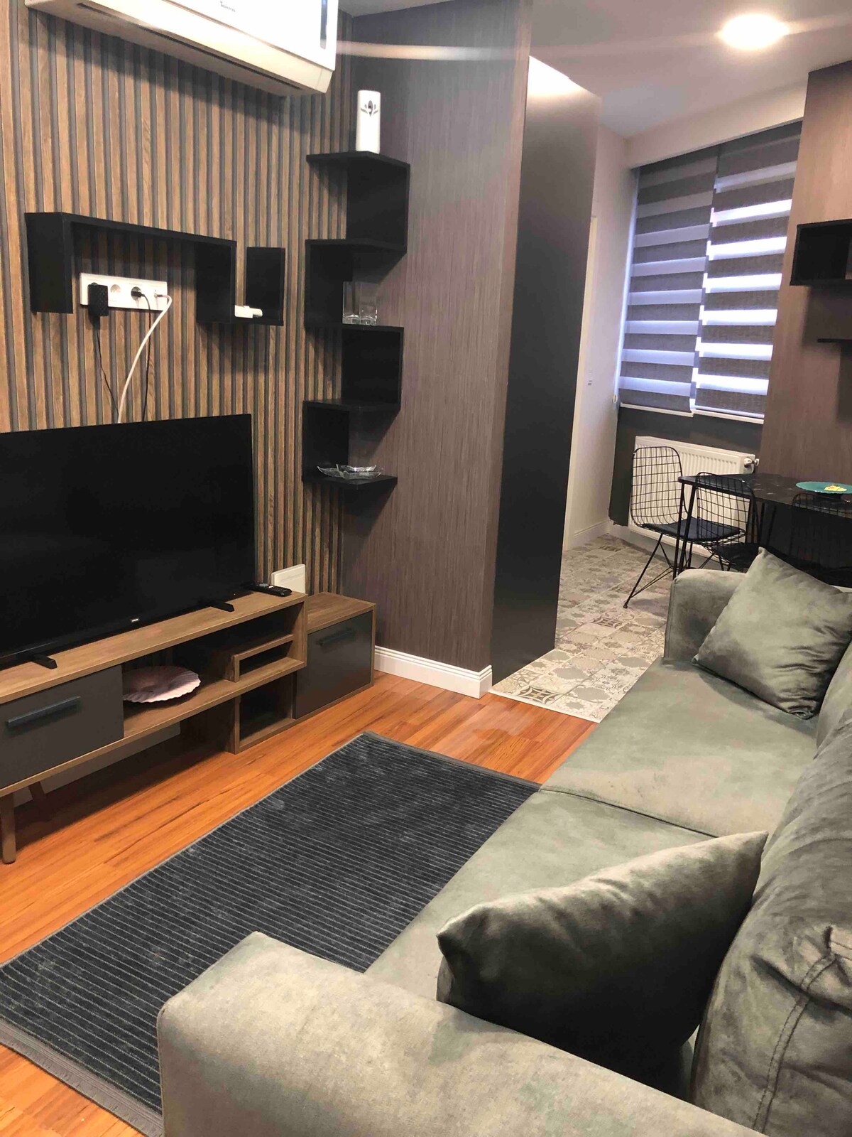 A modern living area features a comfortable green sofa positioned in front of a sleek television stand. Dark wooden accents are complemented by stylish wall shelves. A small dining space with black wire chairs is visible in the background, along with partially open striped blinds allowing natural light.