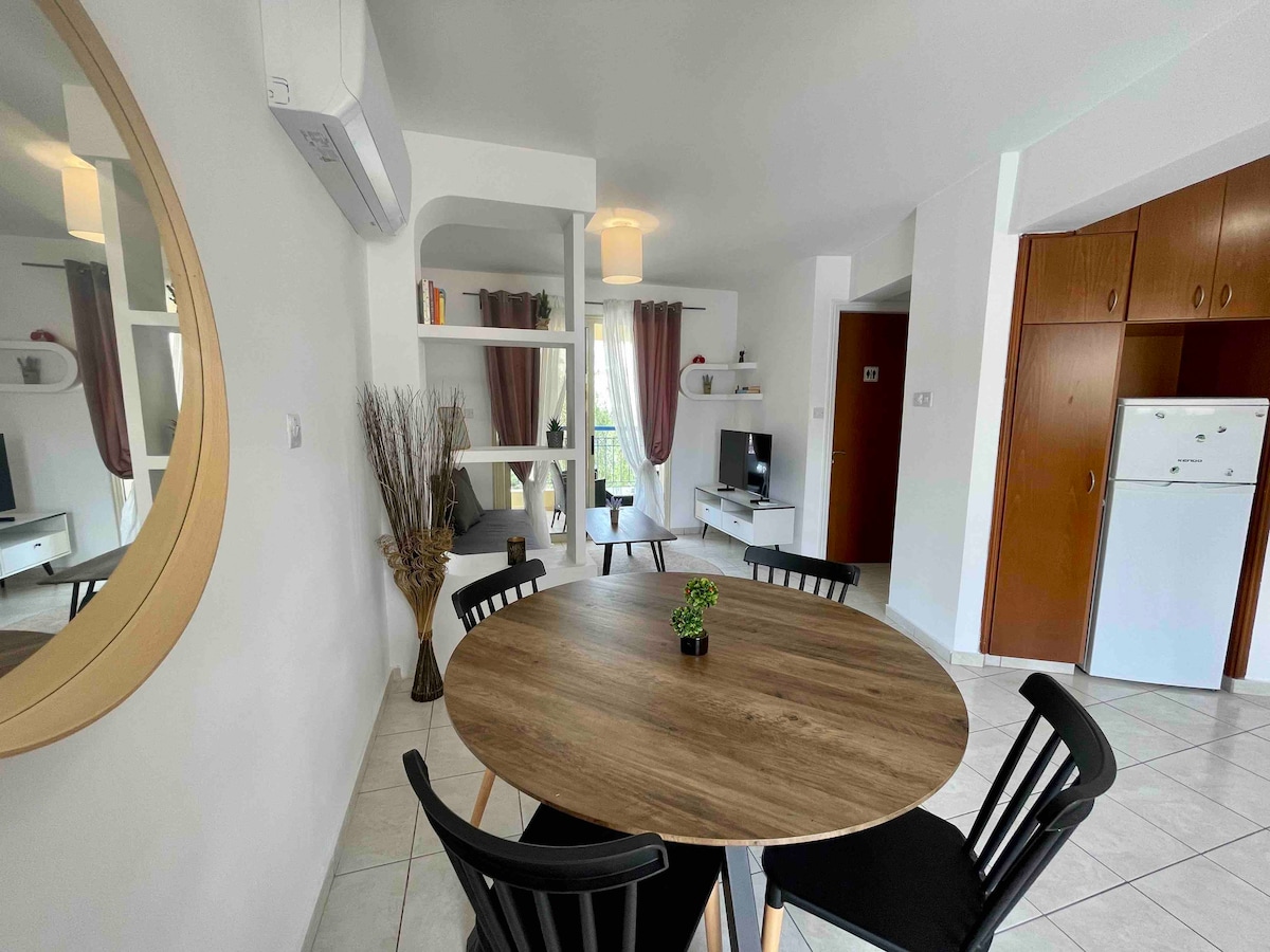 The living area features a circular wooden dining table surrounded by black chairs. A small indoor plant rests on the table. To one side, a comfortable seating arrangement is visible, accompanied by a television on a white console. Natural light floods the space through windows adorned with sheer curtains.