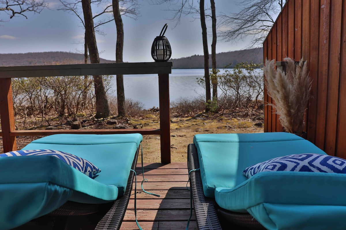 Two lounge chairs are positioned on a wooden deck, offering a view of the tranquil lake surrounded by trees. Decorative pillows add a touch of comfort, while a lantern sits on the railing, creating an inviting outdoor space.