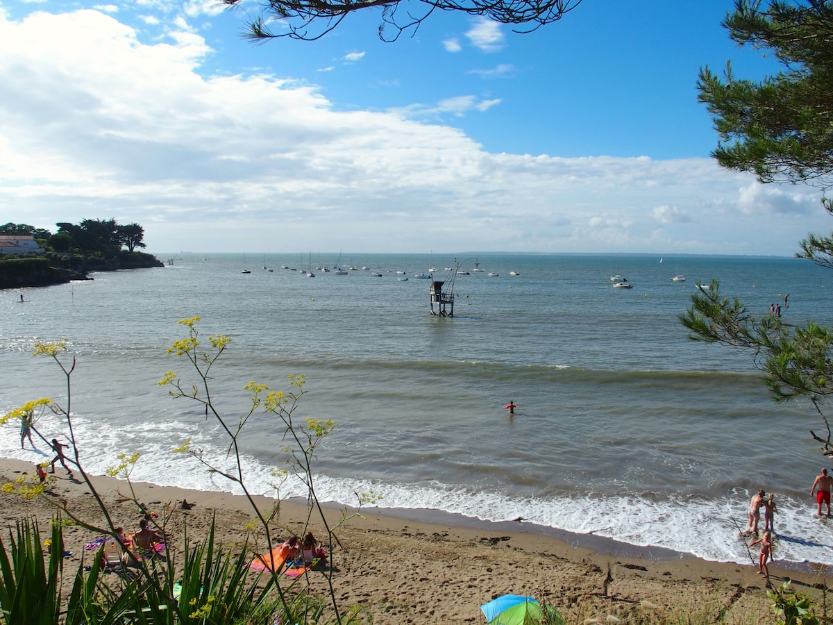 A sandy beach is depicted with gentle waves lapping at the shore. Small boats can be seen on the water, and a wooden platform stands in the distance. Trees frame the scene, providing a natural border against the bright sky.
