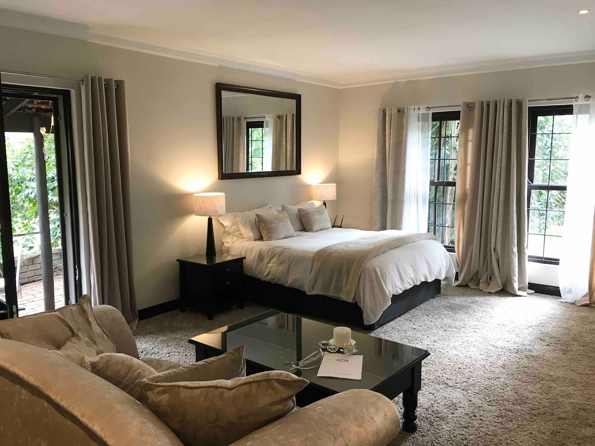 A spacious suite features a comfortable bed, complemented by soft bedding and decorative pillows. Natural light fills the room through large windows draped with sheer curtains, while a plush area rug covers the floor. A stylish coffee table and elegant lamps add functional charm.