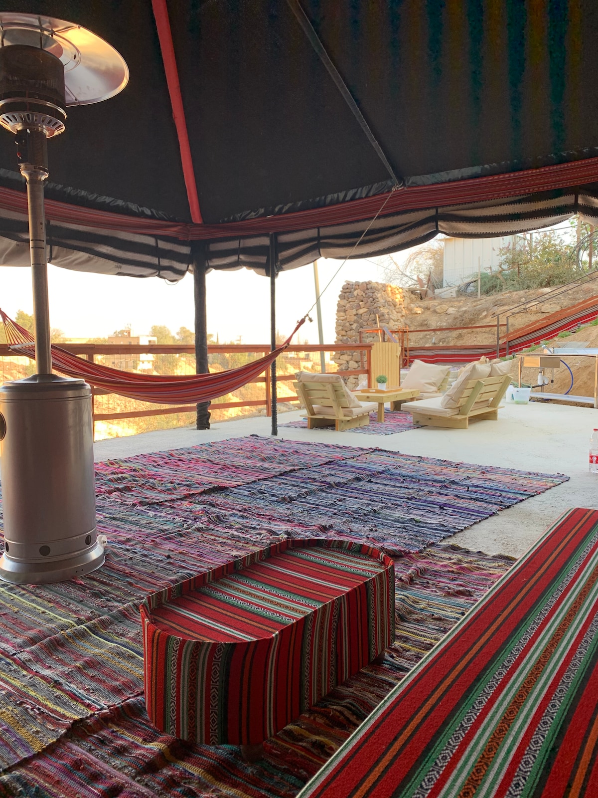 A spacious tent area features colorful woven rugs and a variety of seating options, including two wooden chairs and a patterned low table. Soft hammocks are suspended in the background, with a heater providing warmth. Natural light streams in, creating a bright and inviting space.