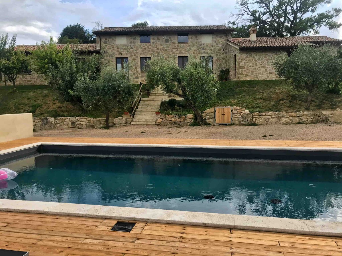 A stone farmhouse sits in the background, surrounded by fruit trees and olive groves. In the foreground, a rectangular swimming pool is bordered by wooden decking. Steps lead up from the pool area to the farmhouse entrance, enhancing the tranquil outdoor space.
