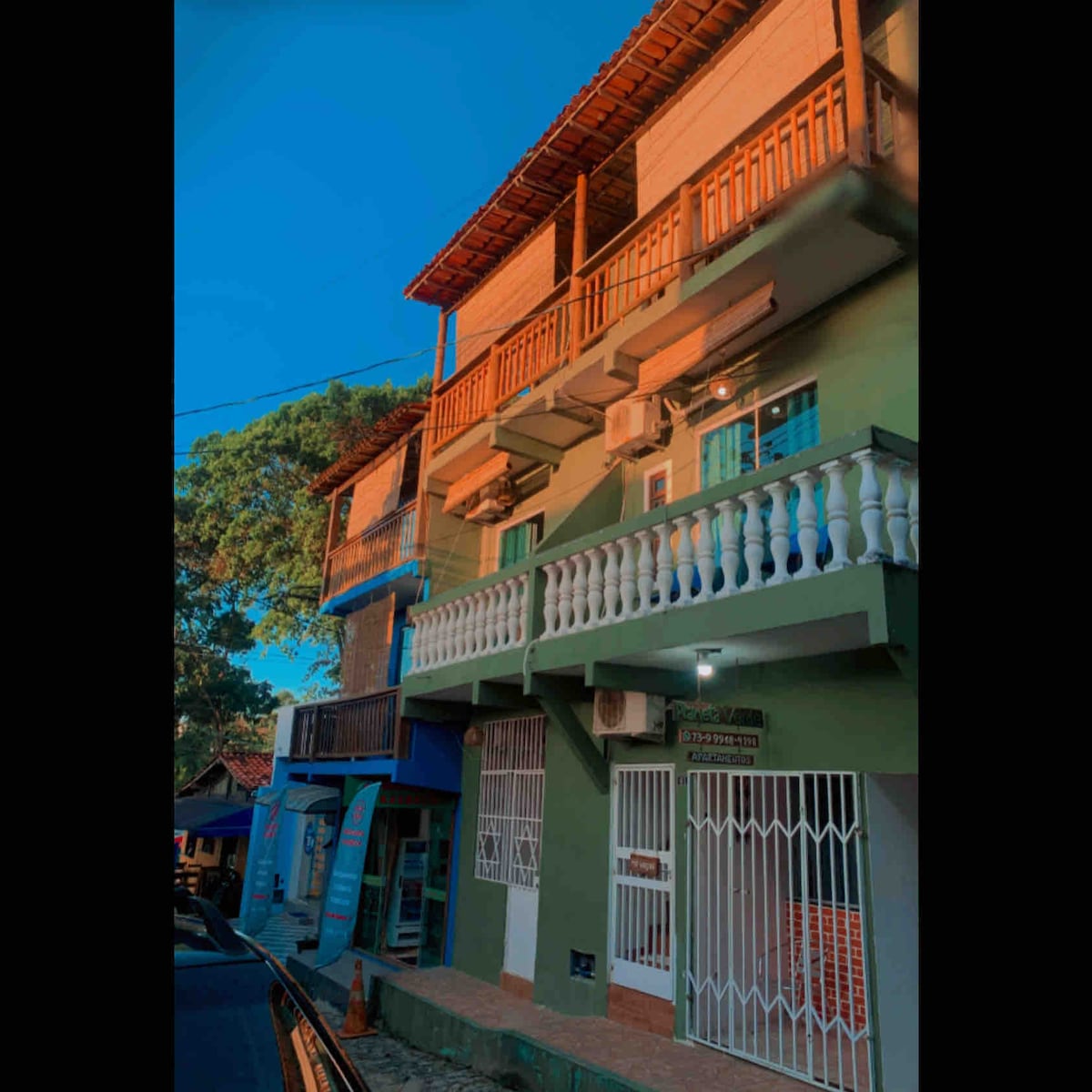 A two-story building is captured in warm evening light, featuring a green exterior with white balconies. Air conditioning units are mounted on the walls, and the ground floor includes a gated entrance. Nearby commercial spaces can be seen, indicating a vibrant area.