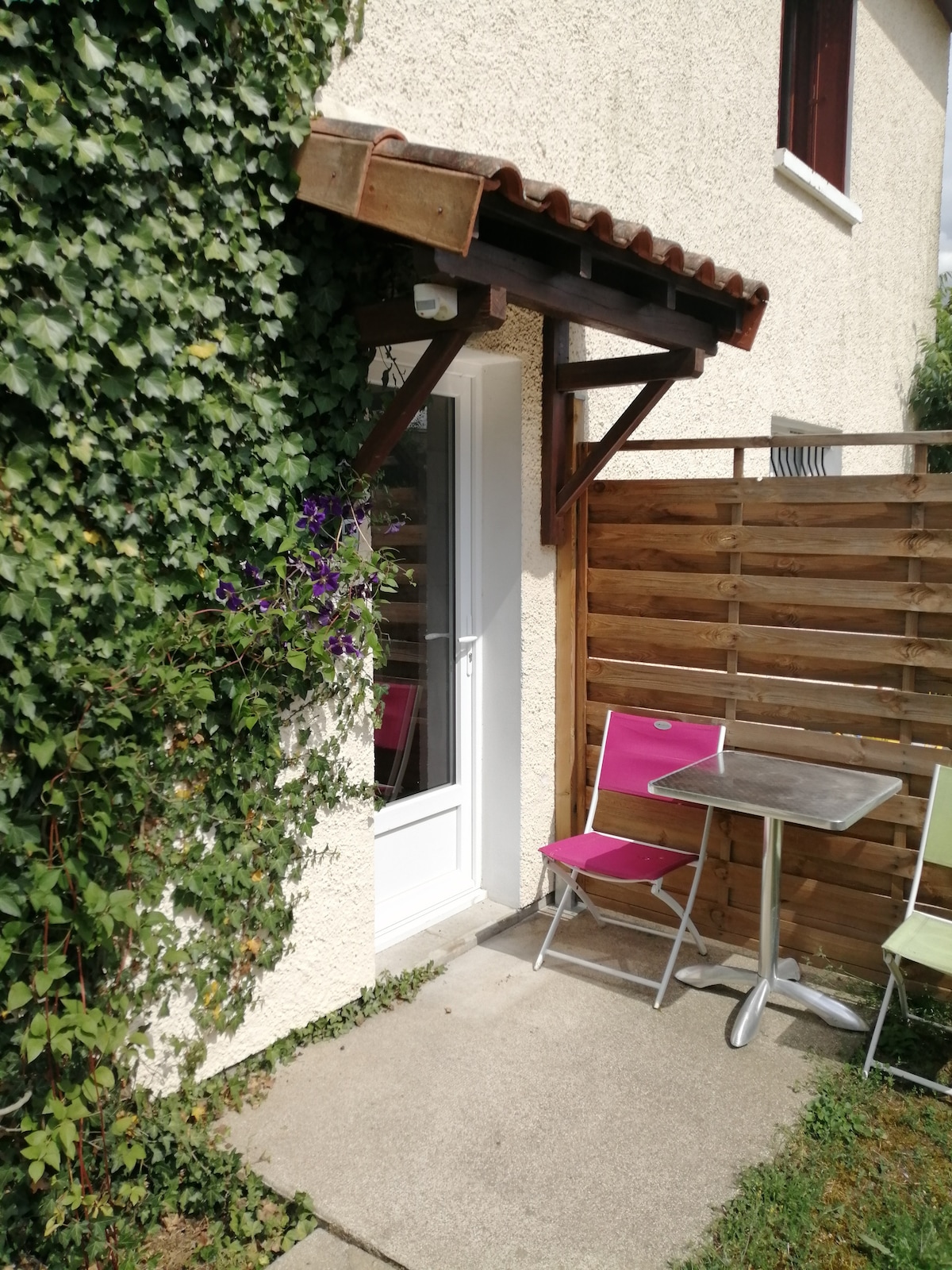 An entrance is framed by lush greenery, featuring an off-white wall and a small awning. A compact table and two colorful folding chairs are positioned on a concrete patio, providing an inviting space for outdoor relaxation.