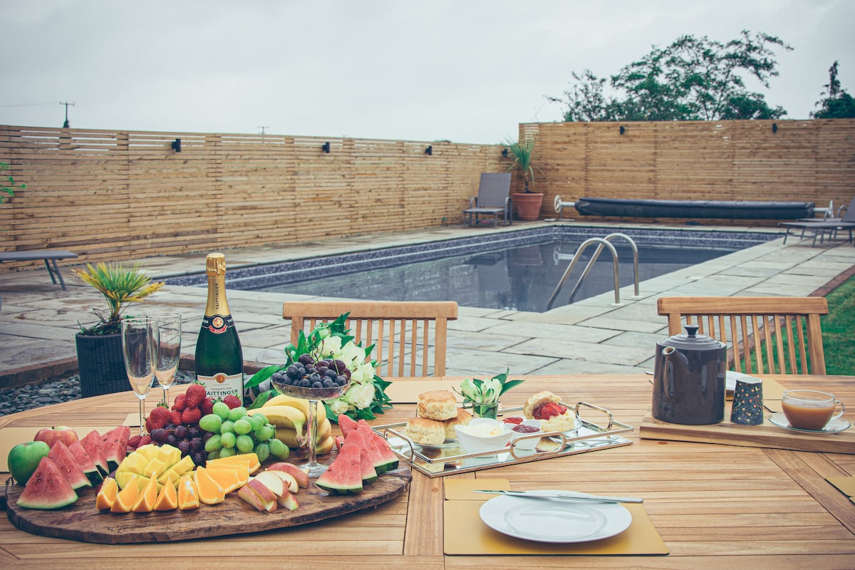 An outdoor dining setup is presented with a wooden table adorned with a fruit platter, champagne, and pastries. In the background, a private swimming pool is visible, surrounded by wooden fencing and greenery, enhancing the serene atmosphere of the space.
