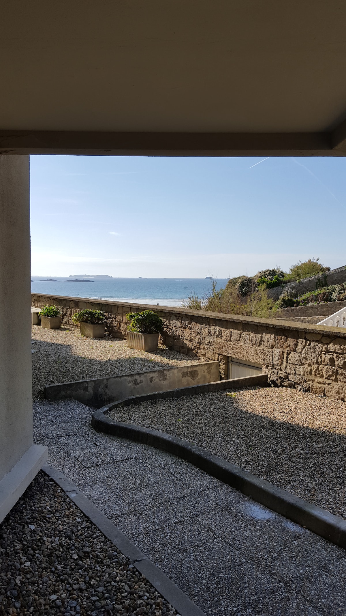 A view of the sea is framed by a stone wall, with gravel paths and potted plants adorning the area. The clear sky and distant islands are visible in the background, reflecting a serene coastal atmosphere.