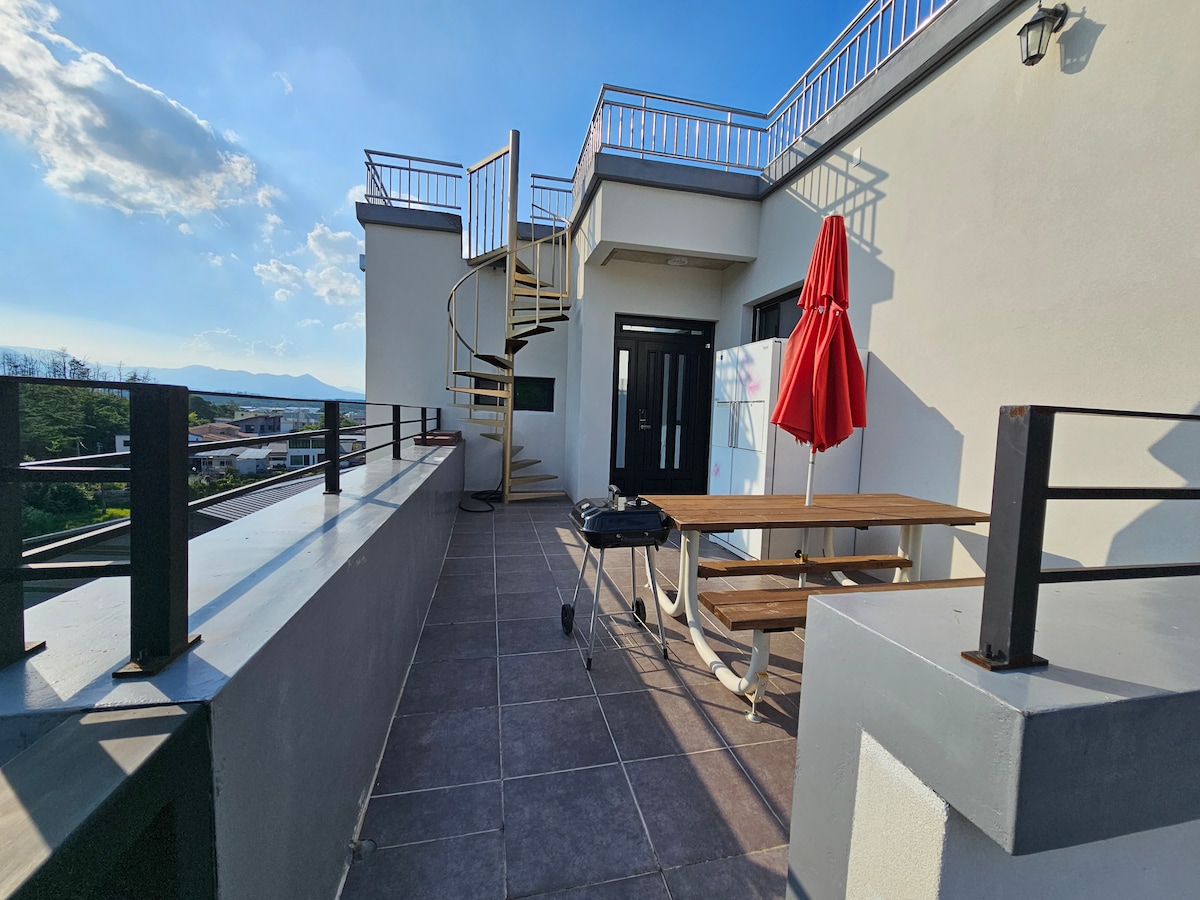 A spacious outdoor terrace features a wooden table with benches and an umbrella. A spiral staircase leads to an upper level. The area is surrounded by low walls, offering panoramic views, while ample sunlight brightens the tile flooring.