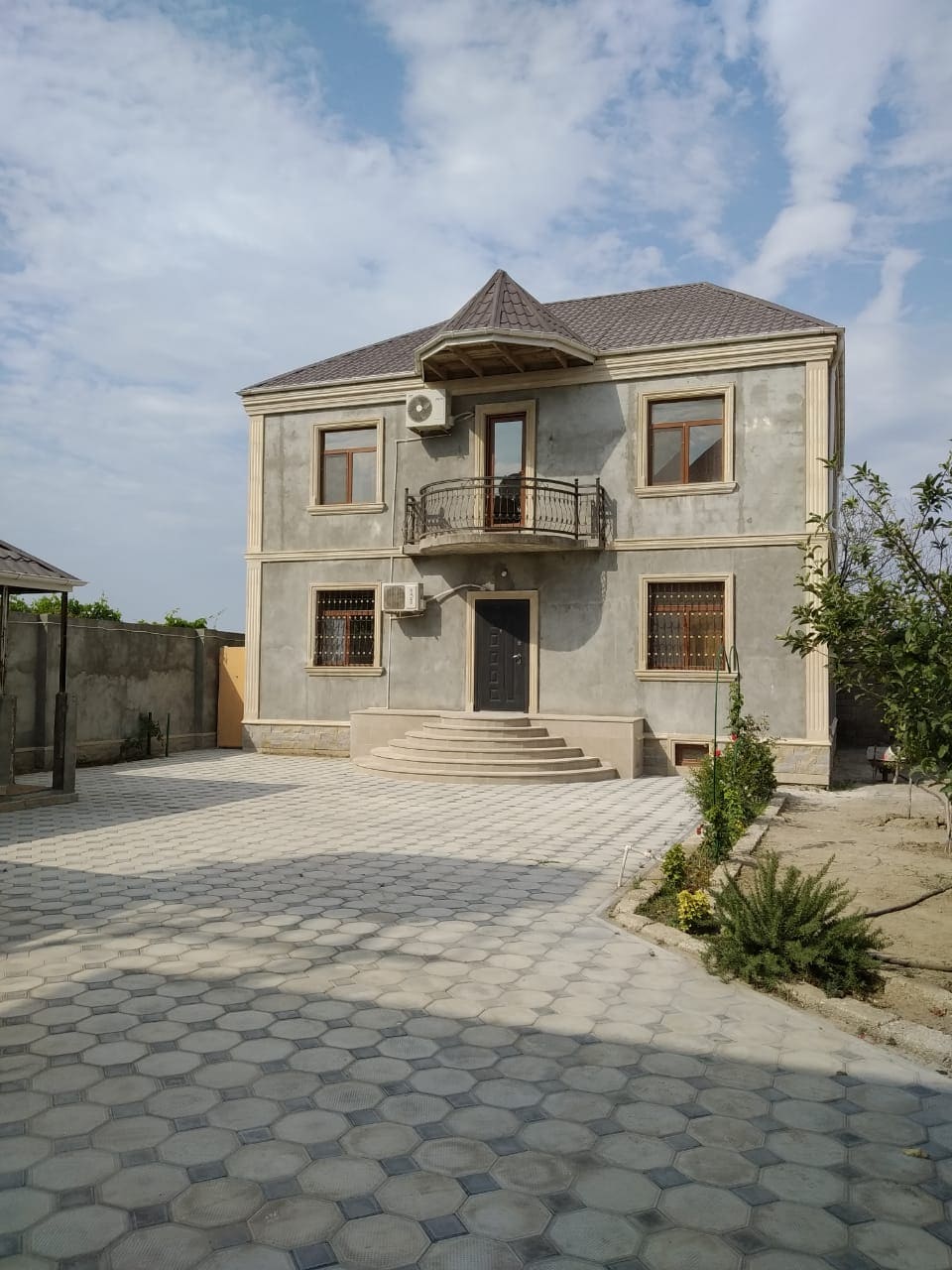 The two-story house features a light gray exterior with decorative stone accents. A covered balcony extends from the second floor, while a solid front door is situated at the center. Paved stone pathways lead through the outdoor area, surrounded by greenery.