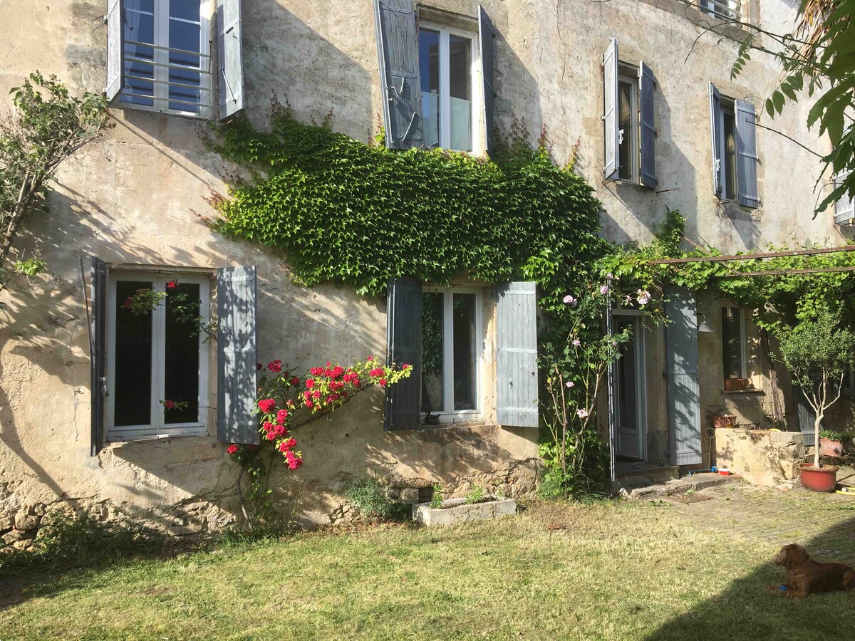 The exterior of the house features rustic stone walls adorned with climbing greenery. Windows framed in wooden shutters open toward the garden, while blooming flowers add vibrant colors to the scene. A well-maintained lawn provides a pleasant area for outdoor activities.