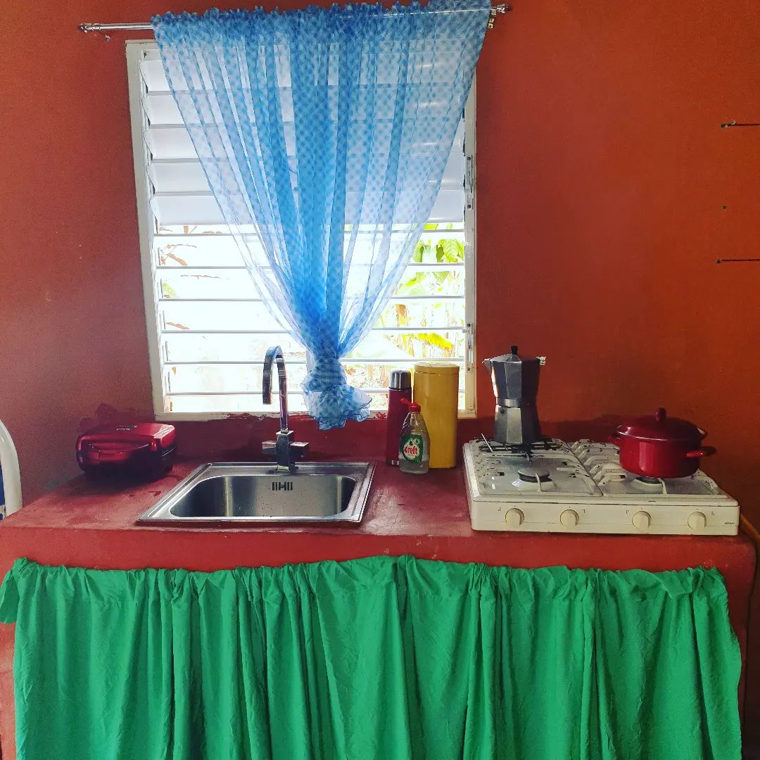 A compact kitchen area features a sink and a gas stove with a pot on top. Bright blue curtains frame a window, allowing natural light to enter. A green fabric covers the countertop, and cooking condiments are displayed nearby, adding a functional touch.