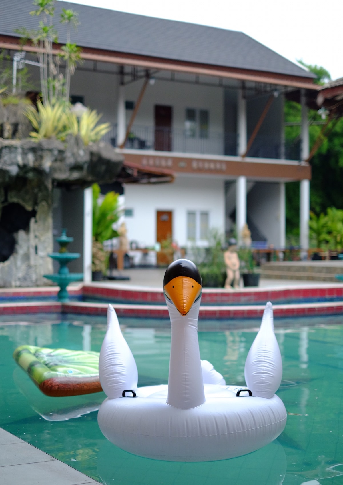 A white swan-shaped float is positioned in the inviting pool, surrounded by lush greenery. The two-story villa is visible in the background, featuring a balcony and multiple windows. A second float appears on the water, complementing the serene atmosphere.