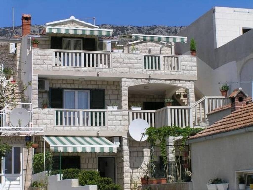 The villa showcases a multi-level structure made from Brač stone, featuring green-and-white striped awnings above several balconies. Potted plants adorn the balcony railings, and satellite dishes are mounted on the exterior walls, contributing to the inviting appearance of the building.