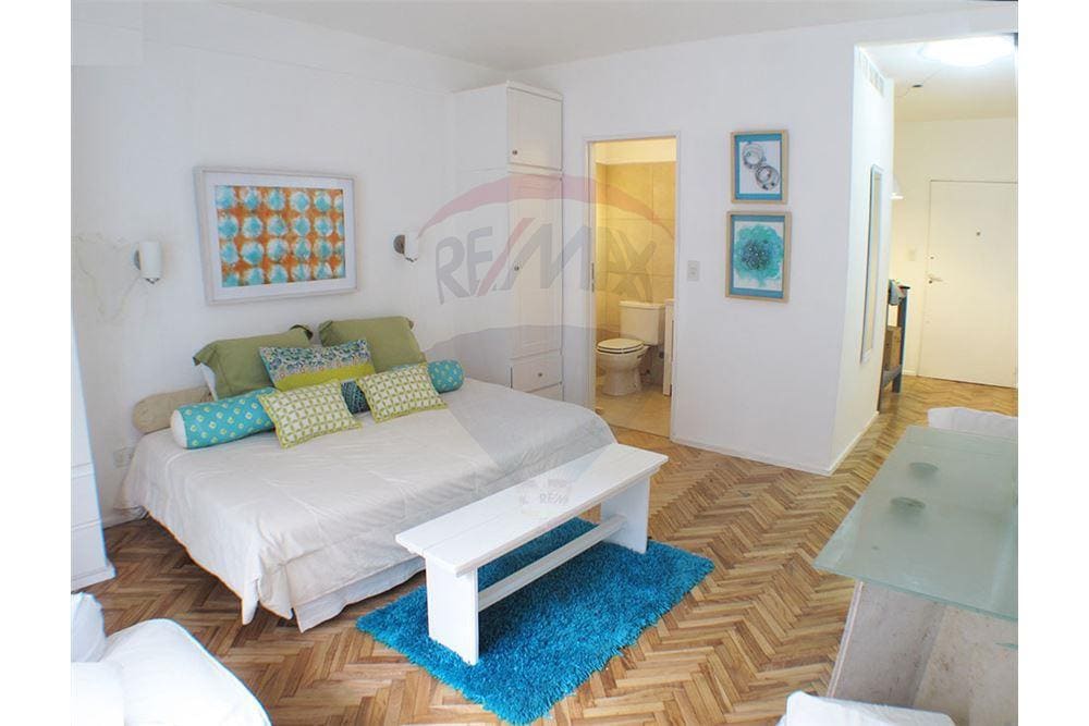 A bright studio apartment is displayed, featuring a spacious bed adorned with colorful pillows and a cozy throw. A white coffee table sits on a plush blue rug, with art pieces on the walls and an open doorway leading to a bathroom.