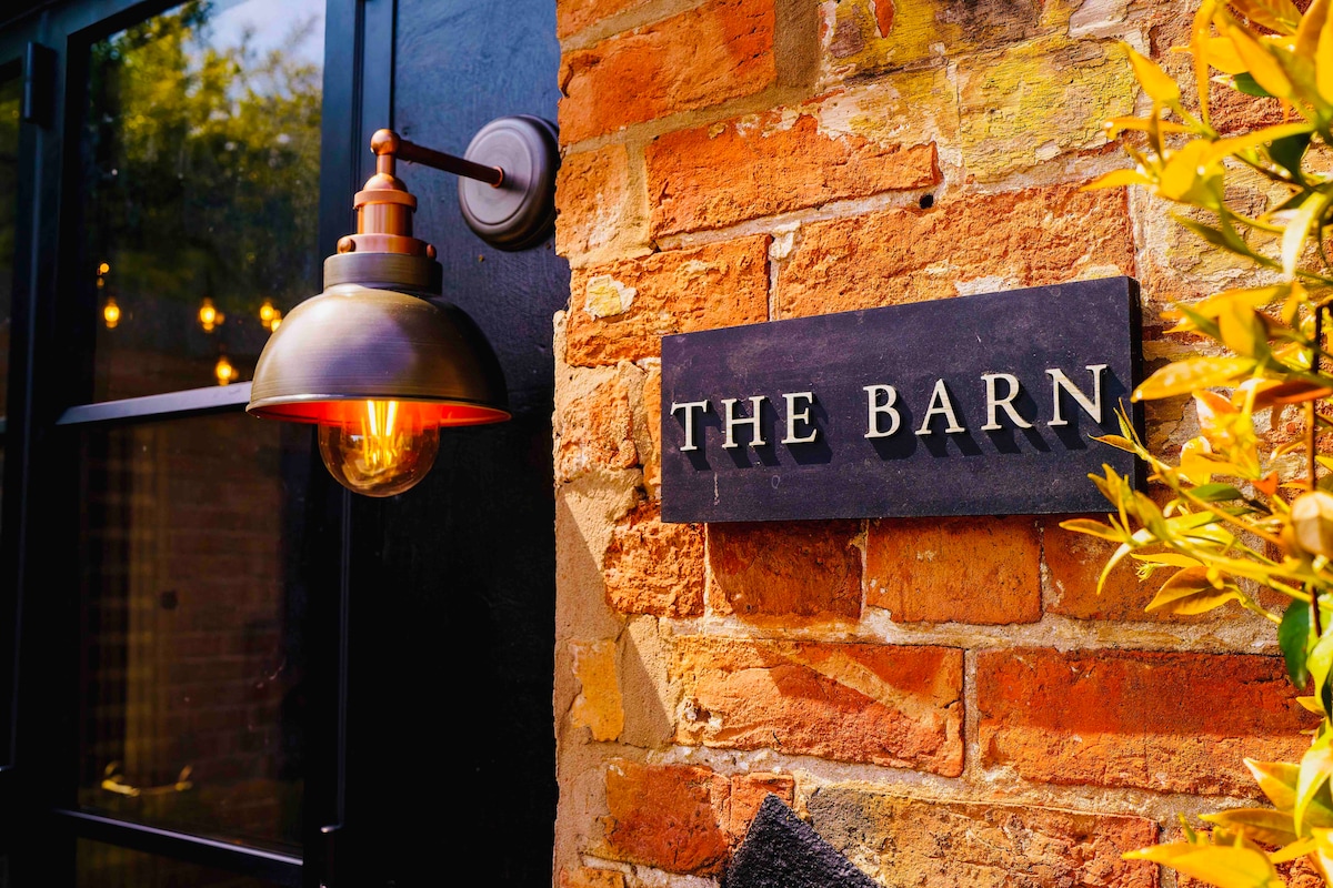 The entrance to The Barn is highlighted by a rustic brick wall and an elegant light fixture. A wooden sign, engraved with 'THE BARN,' is prominently displayed, enhancing the welcoming atmosphere of the retreat.